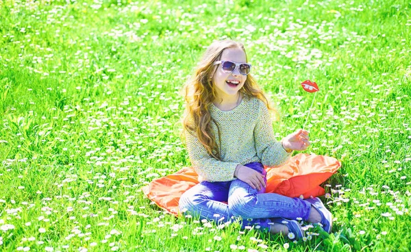 Menina no rosto alegre passar o lazer ao ar livre. Conceito Fashionista. Criança posando com óculos de sol de papelão lábios sorridentes para sessão de fotos no prado. Menina senta-se na grama no grassplot, fundo verde — Fotografia de Stock