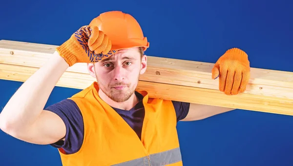 Woodworker concept. Man in protective gloves holds visor of helmet, corrects hard hat on head, blue background. Carpenter, woodworker, labourer, builder carries wooden beam on shoulder — Stock Photo, Image