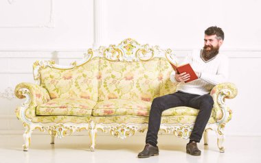 Sakal ve bıyık oturur Barok tarzı kanepe, tutar kitap, beyaz duvar arka plan üzerinde olan adam. Maço yüz kitap okuma konusunda tutkulu gülümseyen üzerinde. Adam zevk ile eski kitap okuma. Okuyucu kavramı