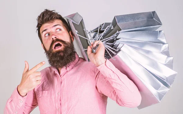 Concepto de venta y descuento. Un tipo de compras en temporada de ventas, apuntando a las bolsas. Hipster en adicto a la cara sorprendido shopaholic. Hombre con barba y bigote lleva montón de bolsas de compras, fondo gris —  Fotos de Stock