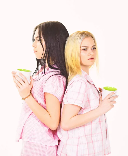 Las hermanas o los mejores amigos en pijama se ponen espalda con espalda. Chicas tomando café por la mañana, fondo blanco aislado. Concepto de café matutino. Rubia y morena en las caras somnolientas sostiene tazas con café — Foto de Stock