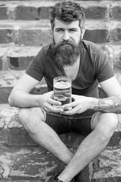 Hipster with long beard looks relaxed. Bearded hipster holds beer mug, drinks beer outdoor. Man with beard and mustache on calm face, stony background, defocused. Craft beer concept