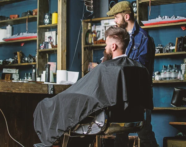 Hipster client got new haircut. Barber with bearded man looking at mirror, barbershop background. Haircut concept. Barber finished trimming. Client and professional master checking result