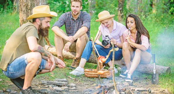 Friends group tourist relaxing near bonfire. Hipster roasting sausage while friends speaking sharing impression and watching photos on camera. Tourists having snack time with roasted over fire food