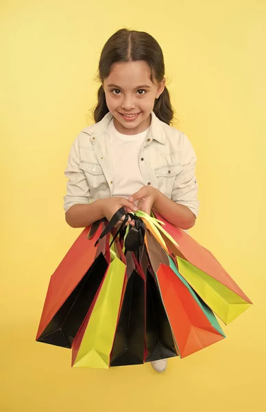 Podívej se na její nákupní balíčky. Dítě roztomilý shopaholic s partou nákupních tašek černé pátek celkový prodej. Vezměte si nákupní balíčky. Dívka nese nákupní tašky žluté pozadí. Dívka ráda nakupuje — Stock fotografie