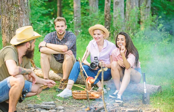 Tourists having snack time with roasted over fire food. Hipster roasting sausage while friends speaking sharing impression and watching photos on camera. Friends group tourist relaxing near bonfire