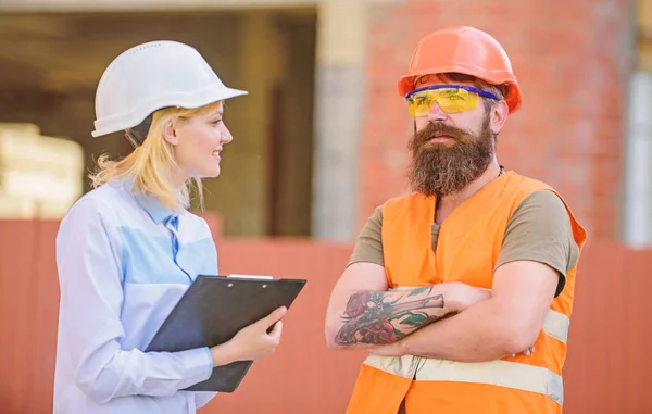 Woman inspector and bearded brutal builder discuss construction progress. Construction project inspecting. Construction site safety inspection. Discuss progress project. Safety inspector concept