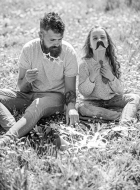Baba ve kızı, grassplot, arka planı yeşil çimenlerin üzerinde oturur. Çocuk ve babası eyeglases ve muastache photo booth öznitelikleri ile poz. Aile zaman kavramı. Aile harcamak açık havada eğlence