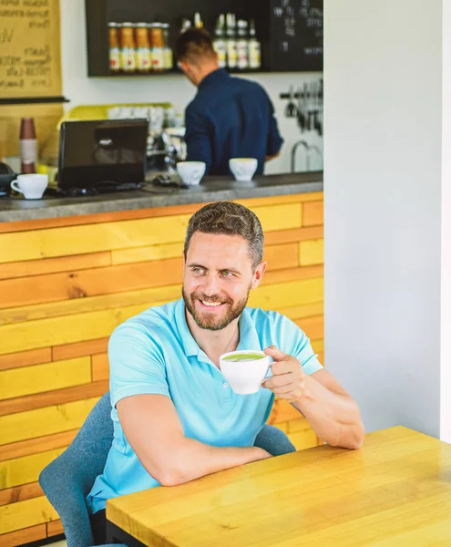 Man bearded guy drinks cappuccino wooden table cafe. Cafe visitor happy smiling face enjoy coffee drink. Improve overall health. Take moment to care about yourself. Coffee drinkers live longer