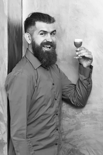 Bearded man, long beard. Brutal caucasian smiling happy hipster with moustache in brown shirt holding alcoholic red shot on golden texture backgroun — Stock Photo, Image
