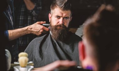 Reflexion of barbers hand with hair clipper trimming nape of client. Hipster bearded client getting hairstyle in front of mirror. Barbershop concept. Barber with hair clipper trimming nape, defocused clipart