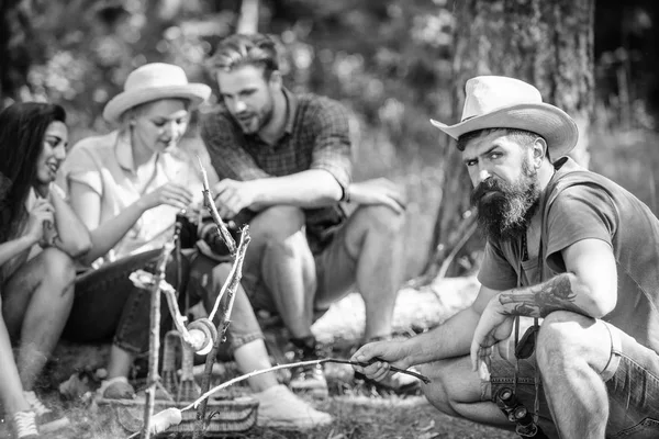 Hipster arrostire salsiccia mentre gli amici si siedono sul log sharing impressione e guardare le foto sulla macchina fotografica. Turisti che fanno merenda con arrosto sul cibo fuoco. Amici gruppo turistico rilassante vicino al falò — Foto Stock