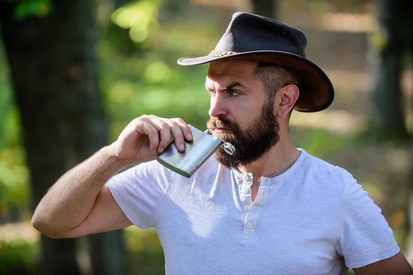 Tipo brutal barbudo vaquero beber alcohol matraz de metal. Hipster con barba beber alcohol naturaleza fondo desenfocado. Camping de senderismo. Concepto vagabundo. Bebida alcohólica. Hombre nómada llevar alcohol con él — Foto de Stock