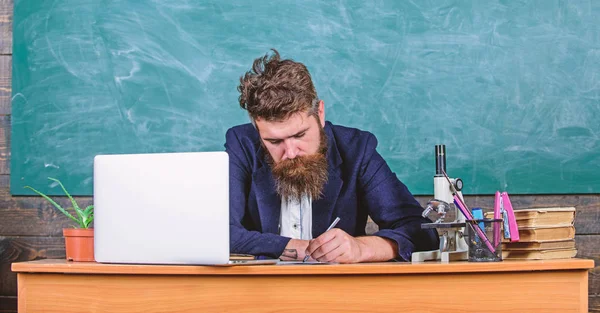 School teacher checking homework or test. School paperwork. Paperwork part of teachers life. Teacher sit desk with laptop. Teacher bearded hipster with eyeglasses sit classroom chalkboard background — Stock Photo, Image