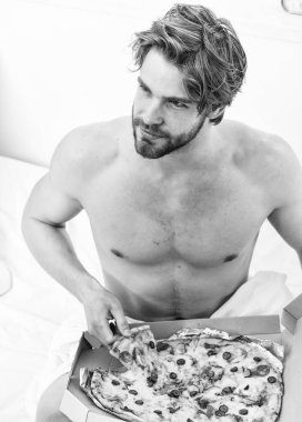 Bir yatağa evde döşeme sırasında tembel kaslı adam yeme pizza portresi. Gömleksiz seksi adam yatakta pizza ile görüntü kırpılmış. Ara diyet kavramı.