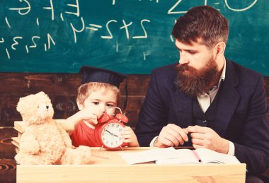 Bireysel ders kavramı. Öğretmen sakallı, sınıfta, arka plan kara tahta küçük oğlu baba öğretir. Öğretmen çocukla konuşurken oğlum, sakin yüz çocuk çalar saat tutar