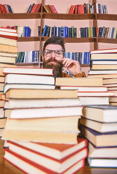 Mann mit nachdenklichem Gesicht zwischen Bücherstapeln in der Bibliothek, Bücherregale im Hintergrund. Lehrer oder Schüler mit Bart tragen Brillen, sitzen mit Büchern am Tisch und sind defokussiert. wissenschaftliches Forschungskonzept — Stockfoto