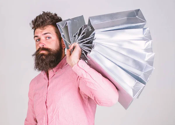 Concepto de compras. Chico de compras en temporada de ventas, lleva un montón de bolsas en el hombro. Hombre con barba y bigote sostiene bolsas de compras, fondo gris. Hipster en serio la cara de compras —  Fotos de Stock