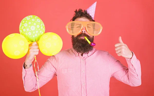 Fiesta de cumpleaños sorpresa con decoración de globos. Hombre barbudo con camisa rosa mostrando el pulgar hacia arriba. Hipster con enormes gafas y silbato de fiesta aislado sobre fondo rojo. Hombre organizando cumpleaños para niños —  Fotos de Stock