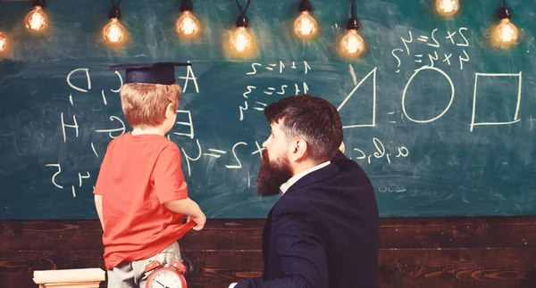 Individual education concept. Teacher and child turned back in the classroom. Boy listening to the explanation about different shapes. Adult pointing at the board while looking at kid. — Stock Photo, Image