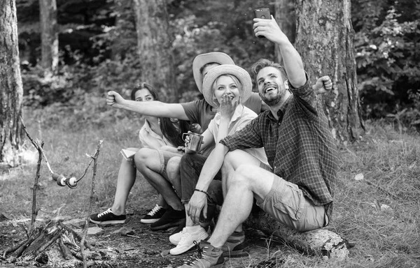 Amici vicino al falò godono di vacanza e cibo arrosto. I turisti si siedono log vicino al falò scattando selfie smartphone foto. Amici in vacanza momento di cattura. Uomo scattare foto vicino a falò natura sfondo — Foto Stock