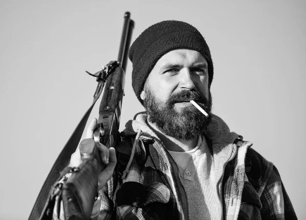 Guy bearded hunter spend leisure hunting and smoking. Hunting masculine hobby concept. Man brutal bearded guy gamekeeper blue sky background. Brutality and masculinity. Hunter with rifle gun close up