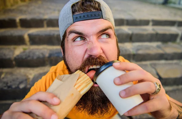 Lanche de bom humor. Conceito de comida de rua. Homem barbudo comer salsicha saborosa e beber copo de papel. Nutrição de estilo de vida urbano. Comida de plástico. hipster despreocupado comer junk food while sit stairs. Cara comer cachorro quente — Fotografia de Stock