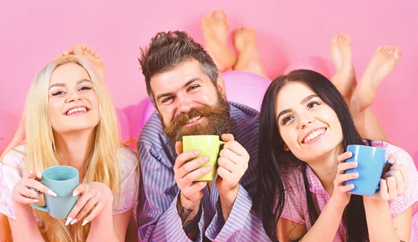 Lovers drinking coffee in bed. Man and women, friends on smiling faces lay, pink background. Man and women in domestic clothes, pajamas. Threesome relax in morning with coffee. Lovers in bed concept