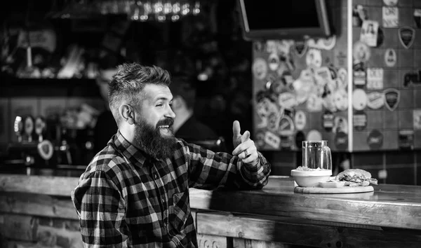 Man with beard drink beer eat burger menu. Enjoy meal in pub. High calorie snack. Hipster relaxing at pub. Pub is relaxing place to have drink and relax. Brutal hipster bearded man sit at bar counter