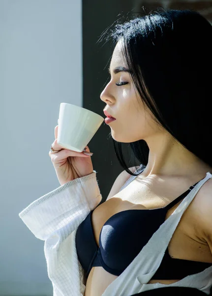 Sip of creamy cappuccino. Morning ritual. Girl sexy breasts drinking coffee near windowsill. Attractive female lingerie relaxing at home. Perfect coffee time. But first coffee. Tranquility and peace