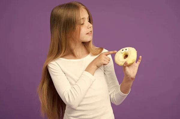Mädchen mit flauschigem Donut. glasierter Bagel mit Augen, kreatives Dessert. Kind isst Sahnehäubchen auf Donut, süßer Zahn Himmel. Kind mit langen Haaren trägt weißen Pullover isoliert auf lila Hintergrund — Stockfoto