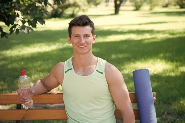 Rumo a um estilo de vida mais saudável. Homem sorridente rosto com tapete de ioga e garrafa de água sentar no banco no parque. Junte-se ao ar livre prática de ioga. Atleta com equipamento de ioga relaxante no parque. Homem escolheu ioga ao ar livre — Fotografia de Stock