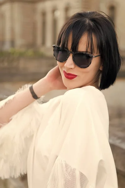 Esconde a emoção dela atrás dos óculos de sol. Menina morena bob penteado parece elegante. Menina senhora elegante com penteado bob ao ar livre arquitetura urbana fundo. Mulher modelo elegante posando ao ar livre — Fotografia de Stock