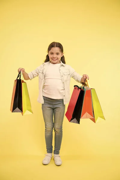 Petite assistante. Enfant fashionista mignon aide à transporter des paquets pendant les achats. Petite vendeuse. Enfant fille heureux sourire visage porte paquets tas fond jaune. Fille shopaholic aime faire du shopping — Photo