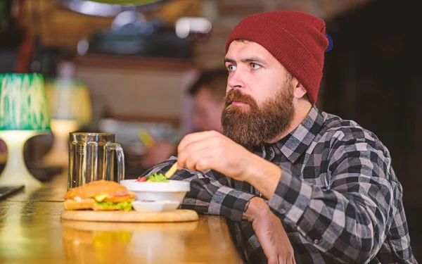 Man with beard eat burger menu. Brutal hipster bearded man sit at bar counter. Cheat meal. High calorie food. Delicious burger concept. Enjoy taste of fresh burger. Hipster hungry man eat burger
