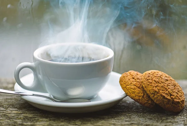 Coffee time on rainy day. Fresh brewed coffee in white cup or mug on windowsill. Wet glass window and cup of hot caffeine beverage. Coffee drink with oat cookies dessert. Enjoying coffee on rainy day