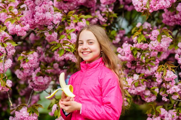 Mała dziewczynka je banana. Dzieciak na różowych kwiatach tła drzewa sakura. Dzieciak cieszący się wiśniową sakurą. Wesołych wakacji. Wiosna w ogrodzie botanicznym. Tak pachnie wiosna. Kwitną kruche kwiaty — Zdjęcie stockowe