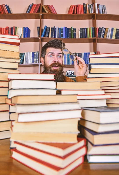 Öğretmen ya da öğrenci sakallı gözlük takmaz, defocused kitaplar ile masada oturur. Bilimsel araştırma kavramı. Adam kazık kitap Kütüphane, kitap rafı arka plan üzerinde arasında sürpriz yüzünde