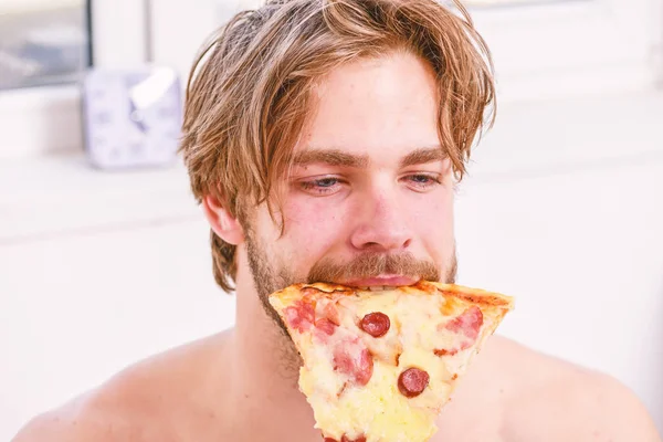 Handsome man holds a piece of pizza in his hands and is about to eat it. Shirtless handsome young man with pizza on bed. Break diet concept. People like pizza.