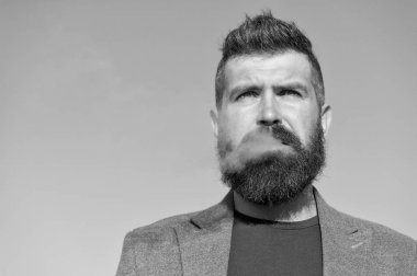 Sakallı ve bıyıklı, mavi gökyüzü arkaplanı tüten bir adam. Sigara ve alışkanlık konsepti. Şık saçlı, düşünceli, konsantre yüzlü, ağzından beyaz duman çıkan bir hippi. Dışarıda sigara içmek.