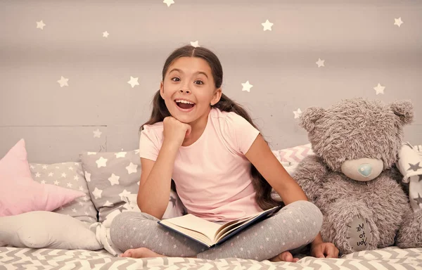 그녀의 좋아하는 동화 같아요. 여자 아이 테 디 베어 책 읽기와 침대를 앉아. 아이 침대에가 서 준비 합니다. 아늑한 침실에서 즐거운 시간입니다. 여자 아이 긴 머리 귀여운 잠 옷 휴식과 장난감 곰 책을 읽고 — 스톡 사진