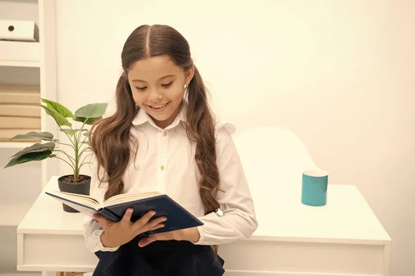 Какая интересная тема. Читаю учебник. Девочка читает книгу, стоя белый интерьер стола. Школьница изучает учебник. Школьная форма для девочек счастливое лицо читает книги. Возбужден знаниями — стоковое фото