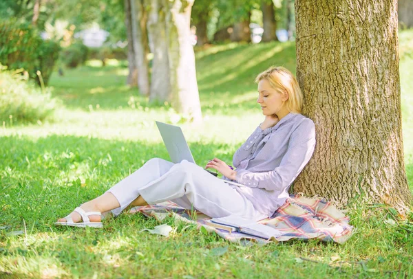Преимущества работы на открытом воздухе. Женщина с ноутбуком работает на открытом воздухе опираясь на ствол дерева. Образовательные технологии и интернет-концепция. Девушка, работающая с ноутбуком в парке, сидит на траве. Природный офис — стоковое фото