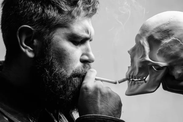 Habit to smoke tobacco bring harm to your body. Smoking cause health damage and death. Man smoking cigarette near human skull symbol death. Nicotine destroy health. Harmful habits. Smoking is harmful