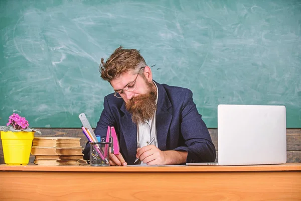 El papeleo forma parte de la vida de los profesores. Profesor barbudo hipster con gafas se sientan en el fondo de pizarra aula. Profesora de escuela revisando tarea o prueba. Profesor sentarse escritorio con portátil. Revisar tarea —  Fotos de Stock