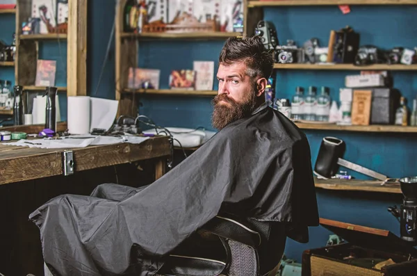 L'uomo con la barba ricoperta di mantello nero siede in sedia parrucchieri davanti allo specchio. Hipster con barba aspetta barbiere e taglio di capelli. Uomo con barba cliente del barbiere hipster. Concetto di barbiere — Foto Stock