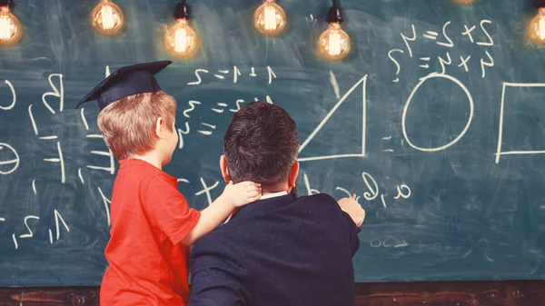 Αγόρι, παιδί στο μεταπτυχιακό ΚΓΠ ακρόασης δάσκαλος, chalkboard σε φόντο, πίσω όψη. Δάσκαλος με γένια, ο πατέρας διδάσκει μικρό γιο στην τάξη, chalkboard σε φόντο. Δάσκαλος αγαπημένες έννοια — Φωτογραφία Αρχείου