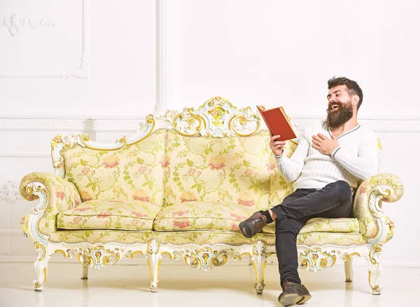 Un tipo leyendo un libro viejo con placer. Hombre con barba y bigote se sienta en el sofá de estilo barroco, sostiene libro, fondo blanco de la pared. Macho en el libro de lectura de caras de risa. Concepto de literatura humorística —  Fotos de Stock