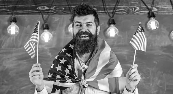 American teacher waves with american flags. Man with beard and mustache on happy face holds flags of USA, in classroom, chalkboard on background. Patriotic education concept. Student exchange program