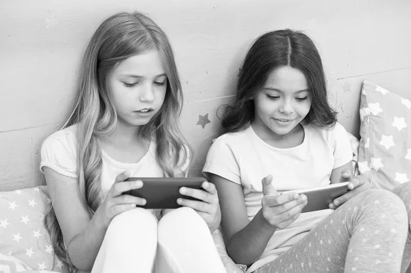 子供の楽しみのためのアプリケーション。インターネットサーフィンと不在親のアドバイス。スマートフォンのインターネットアクセス。女の子の姉妹はスマートフォンで忙しいパジャマを着ています。パジャマの子供たちはスマートフォンと対話します — ストック写真